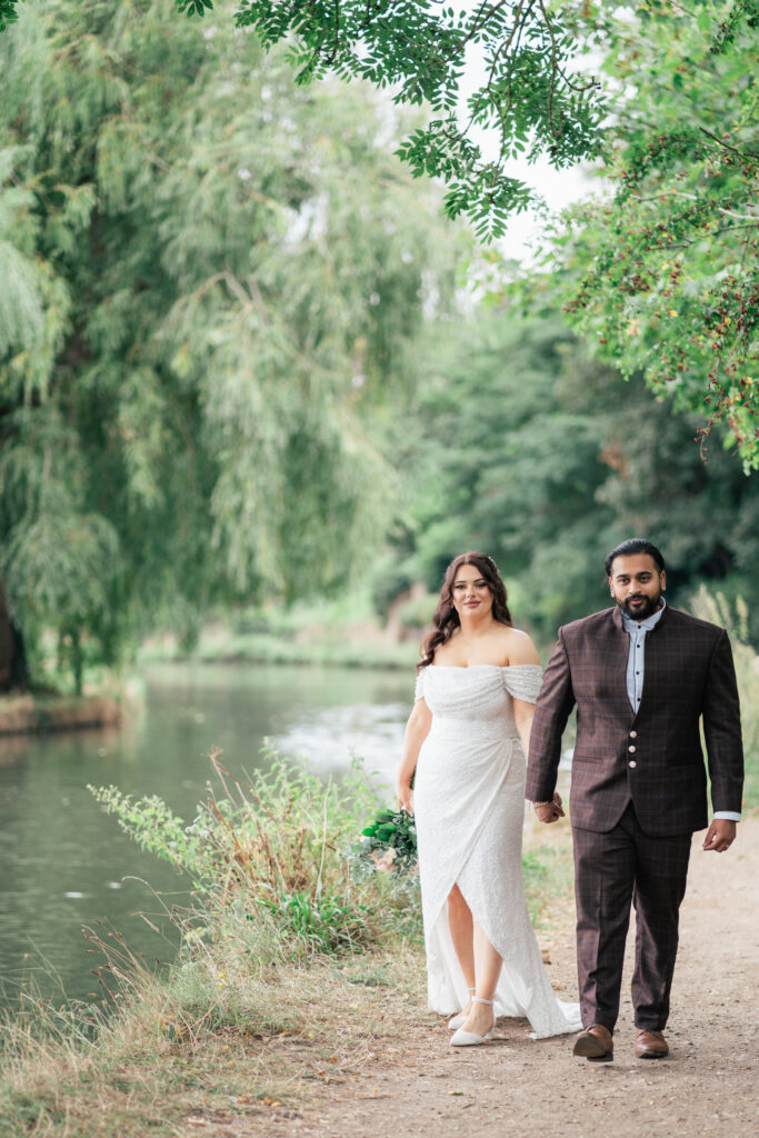 A couple walks hand-in-hand along a riverside path, captured by a Guildford Registry Office photographer. The woman wears an off-shoulder white dress and glasses; the man is in a dark plaid suit amidst green trees and calm water.